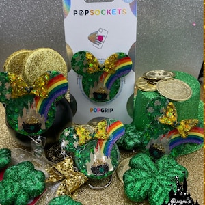 May include: A green and gold glitter resin charm with a rainbow, a pot of gold, and a castle. The charm is shaped like Mickey Mouse ears and has a gold bow. The charm is on a gold background with other charms and a sign that says "Grandma's Main Street Designs".