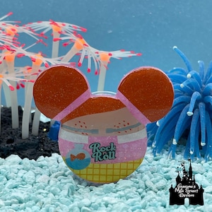 May include: A colorful, glittery pin in the shape of a mouse head with orange ears and a pink and white body. The pin has the words "Rock Roll" and a fish graphic. The pin is set against a blue background with decorative coral.