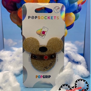 Puede incluir: Una agarradera para teléfono PopSocket de brillo dorado con una banda de cuero marrón y la palabra "SQUIRREL!" impresa en ella. El PopSocket está empaquetado en una tarjeta blanca con el logotipo de PopSockets y las palabras "PHONE GRIP &" y "NICE TO GRIP".