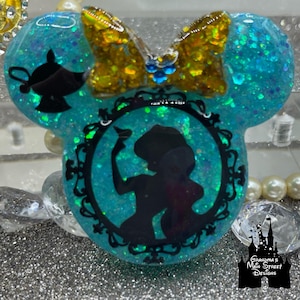 Könnte beinhalten: Ein türkisblaues Mickey-Maus-Ohrenband aus Harz mit einer Schleife aus goldenen Glitzer und einer Silhouette von Jasmin aus Aladdin in einem schwarzen, verzierten Rahmen.