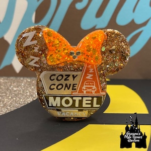 Może przedstawiać: Błyszcząca, złota przypinka w kształcie uszu Myszki Miki z pomarańczową kokardą. Przypinka przedstawia znak Cozy Cone Motel i stożkowy wzór z samochodem. Tekst "ZZZ" jest widoczny na uszach i stożku.