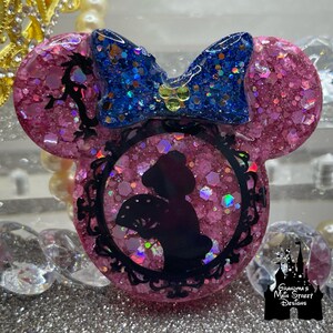 Könnte beinhalten: Rosa und blaue Glitzer-Harz-Mickey-Mouse-Ohren mit einer schwarzen Silhouette einer Frau, die einen Fächer in einem dekorativen Rahmen hält.