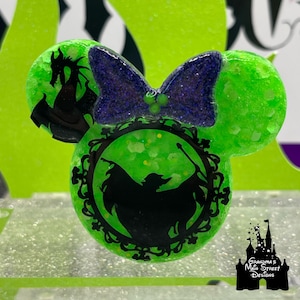 May include: A green, glittery mouse-ear shaped ornament with a purple bow. The ornament features a black silhouette of a dragon on one ear and a witch in a decorative frame on the other. The logo "Grandma's Main Street Designs" is in the bottom right.