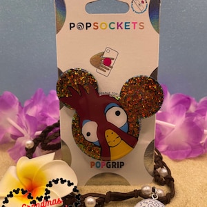 Puede incluir: Un PopSocket para teléfono de color rojo y dorado con purpurina que presenta un gallo de dibujos animados con ojos grandes. El PopSocket está empaquetado sobre un fondo plateado con el texto "PopSockets" y "PopGrip".