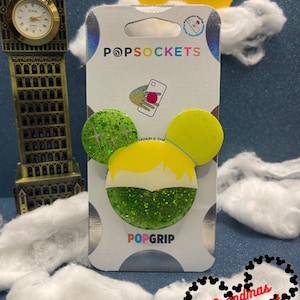 May include: Green and yellow glitter phone grip with a cartoon character's face. The grip is shaped like Mickey Mouse ears. The packaging says "PopSockets" and "PopGrip".