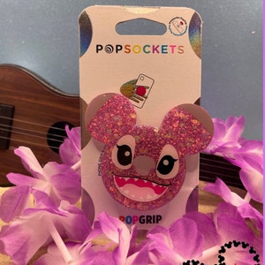May include: Pink glitter phone grip with a cartoon Stitch character design. The grip is packaged on a white card with the words "PopSockets" and "PopGrip" printed on it. The card also features a small red heart with the text "Grandmas Main Street Designs" printed on it.