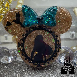 Könnte beinhalten: Ein goldfarbenes Glitzer-Harz-Accessoire in Minnie-Maus-Form mit einer teal-farbenen Glitzer-Schleife. Das Design umfasst eine schwarze Silhouette einer Frau in einem dekorativen Rahmen und einen Kolibri. Der Text "Grandma's Main Street Designs" ist vorhanden.