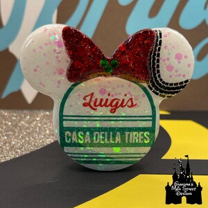 Puede incluir: Un accesorio blanco y brillante con forma de oreja de ratón, con un lazo rojo con purpurina y un diseño de neumático. El accesorio presenta el texto "Luigis CASA DELLA TIRES" en rojo y verde. Un diseño de Grandma's Main Street Designs.