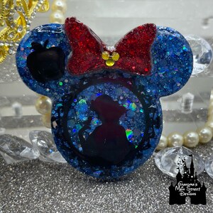 Könnte beinhalten: Blaue und rote Glitzer-Harz-Mickey-Mouse-Ohren mit einer Silhouette von Schneewittchen in einem Rahmen. Die Schleife ist rot mit gelben Glitzerakzenten. Der Text "Grandma's Main Street Designs" ist in der rechten unteren Ecke sichtbar.