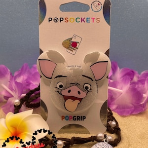 Puede incluir: Un PopSocket blanco y rosa con purpurina que representa a Pua de la película de Disney Moana. El PopSocket está en una tarjeta blanca con las palabras "PopSockets" y "PopGrip" impresas en ella. El PopSocket está rodeado por un cordón marrón con cuentas blancas y un dije de concha azul y blanca.