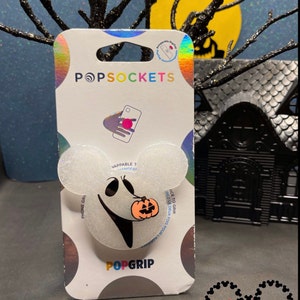 May include: A white PopSocket phone grip with a glittery finish featuring a black and white ghost holding a jack-o-lantern. The PopSocket is packaged in a clear plastic bag with the PopSockets logo and the words "PopGrip" and "VAPPABLE T" printed on it.