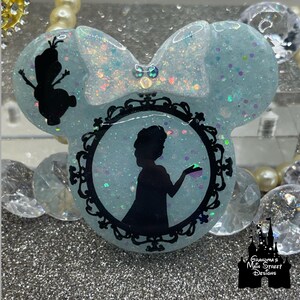 Könnte beinhalten: Ein hellblaues Resin-Mickey-Mouse-Ohrenband mit einer schwarzen Silhouette einer Prinzessin, die eine Tasse in einem schwarzen, verzierten Rahmen hält. Das Stirnband ist mit schimmerndem Glitzer und einer schwarzen Schleife verziert. Der Text "Grandma's Main Street Designs" ist in der unteren rechten Ecke sichtbar.