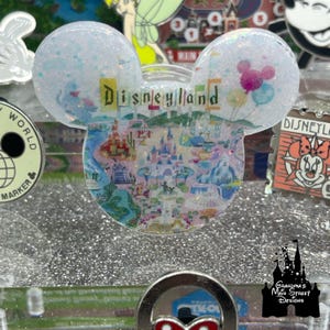 以下が含まれることがあります： 中央に「Disneyland」の文字が入った、透明でキラキラ光るミッキーマウスの耳の形をした装飾品。耳は青いグリッターで満たされ、ディズニーランドの地図とカラフルな風船が描かれています。