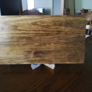 May include: A rectangular wooden sign with a natural wood grain finish. The sign is stained a dark brown color and has a smooth surface. The sign is sitting on a white stand.