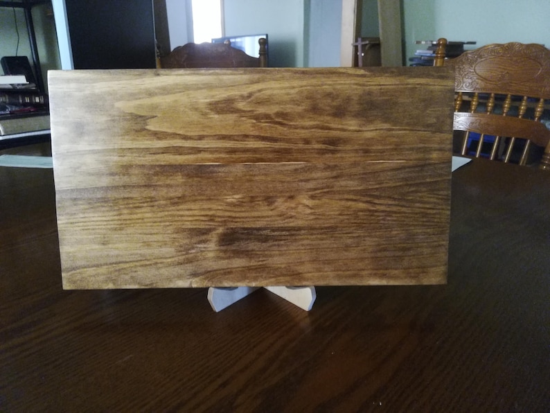May include: A rectangular wooden sign with a natural wood grain finish. The sign is stained a dark brown color and has a smooth surface. The sign is sitting on a white stand.