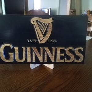 May include: A wooden sign with a black background and a carved Guinness logo. The logo features a harp with the text "ESTD 1759" and "GUINNESS" below it.