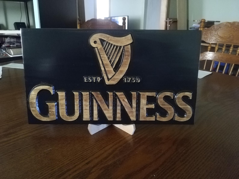 May include: A wooden sign with a black background and a carved Guinness logo. The logo features a harp with the text "ESTD 1759" and "GUINNESS" below it.