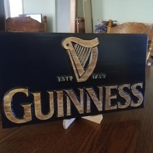 May include: A wooden sign with a black background and a gold Guinness logo. The logo features a harp and the text "ESTD 1759 GUINNESS".