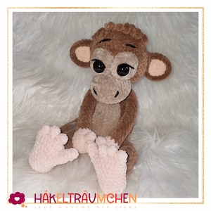 May include: A crocheted brown monkey toy with pink feet sitting on a white fluffy surface. The monkey has large black eyes and a brown nose. The text "HÄKELTRÄUMCHEN JEDE MASCHE MIT LIEBE" is at the bottom of the image.