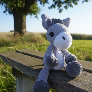 May include: A gray and white crocheted donkey plush toy sitting on a wooden bench. The donkey has a white face with black eyes and a gray body.