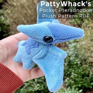 May include: A light blue plush pterodactyl toy with a darker blue beak and wings. The stuffed animal has a large black eye and is labelled "PattyWhack's Pocket Pteradnodon Plush Pattern PDF". The toy is made of soft, fuzzy material.