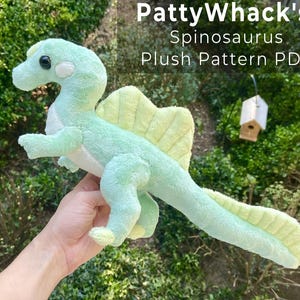 Può includere: Un peluche a forma di dinosauro Spinosaurus di colore verde chiaro e giallo pallido con una superficie morbida e testurizzata, occhi scuri e una pinna dorsale prominente. Il testo PattyWhack's Spinosaurus Plush Pattern PDF è visibile.