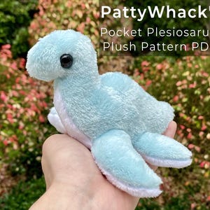 May include: A light blue plush plesiosaurus toy with a black eye is held in a hand. The toy has a soft, textured appearance and white accents on its flippers and belly. Text in the image reads "PattyWhack's Pocket Plesiosaurus Plush Pattern PDF."