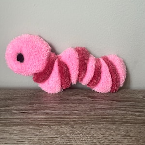 May include: A pink and red striped caterpillar-shaped plush toy. The caterpillar has a large, round head with a black eye and a segmented body with alternating pink and red stripes. The toy is made of a soft, fuzzy material.