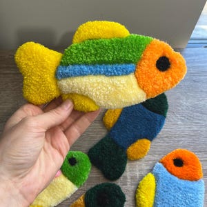 K&ouml;nnte beinhalten: Handgefertigte, fischf&ouml;rmige Dekorationen in verschiedenen Farben. Die Fische sind gelb, gr&uuml;n, blau und orange und bestehen aus einem strukturierten Material. Jeder Fisch hat ein schwarzes Auge. Diese Artikel sind wahrscheinlich f&uuml;r die Inneneinrichtung gedacht.
