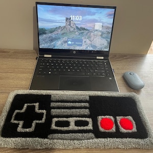 May include: A rectangular tufted rug featuring a black and gray retro video game controller design with two bright red circular buttons. The rug is positioned in front of a silver laptop computer and a small light blue wireless computer mouse.