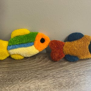 K&ouml;nnte beinhalten: Zwei farbenfrohe, pl&uuml;schige Fischdekorationen. Ein Fisch hat gelbe, gr&uuml;ne, blaue und orange Farbt&ouml;ne mit einem schwarzen Auge. Der andere Fisch hat orange, gelbe und gr&uuml;ne T&ouml;ne. Diese dekorativen Fische sind wahrscheinlich handgefertigt.