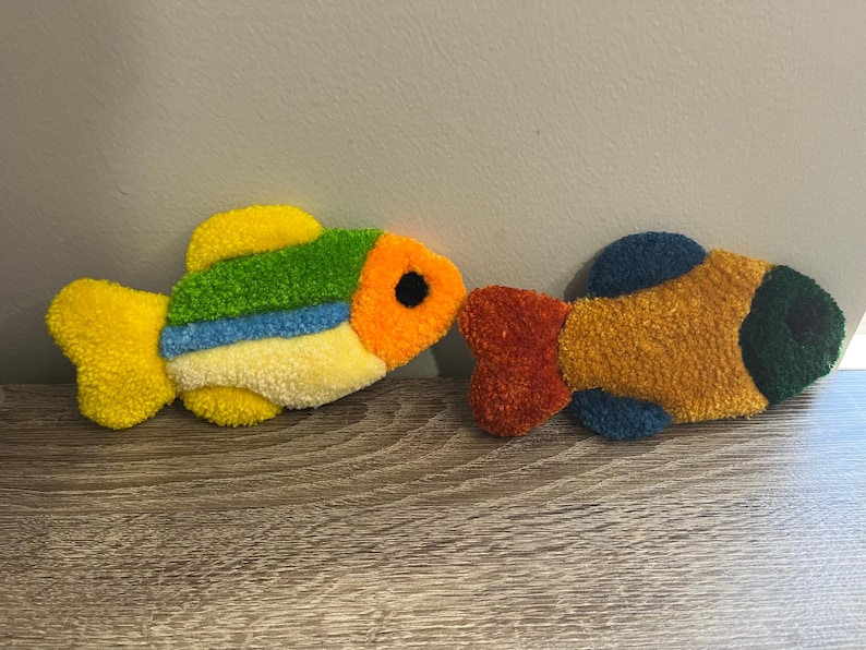 K&ouml;nnte beinhalten: Zwei farbenfrohe, pl&uuml;schige Fischdekorationen. Ein Fisch hat gelbe, gr&uuml;ne, blaue und orange Farbt&ouml;ne mit einem schwarzen Auge. Der andere Fisch hat orange, gelbe und gr&uuml;ne T&ouml;ne. Diese dekorativen Fische sind wahrscheinlich handgefertigt.