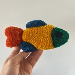 K&ouml;nnte beinhalten: Ein farbenfrohes, strukturiertes Fischornament. Der Fisch hat einen gelben K&ouml;rper, einen roten Schwanz, einen gr&uuml;nen Kopf und blaue Flossen. Das Objekt ist wahrscheinlich handgefertigt und hat ein weiches, pl&uuml;schiges Aussehen.