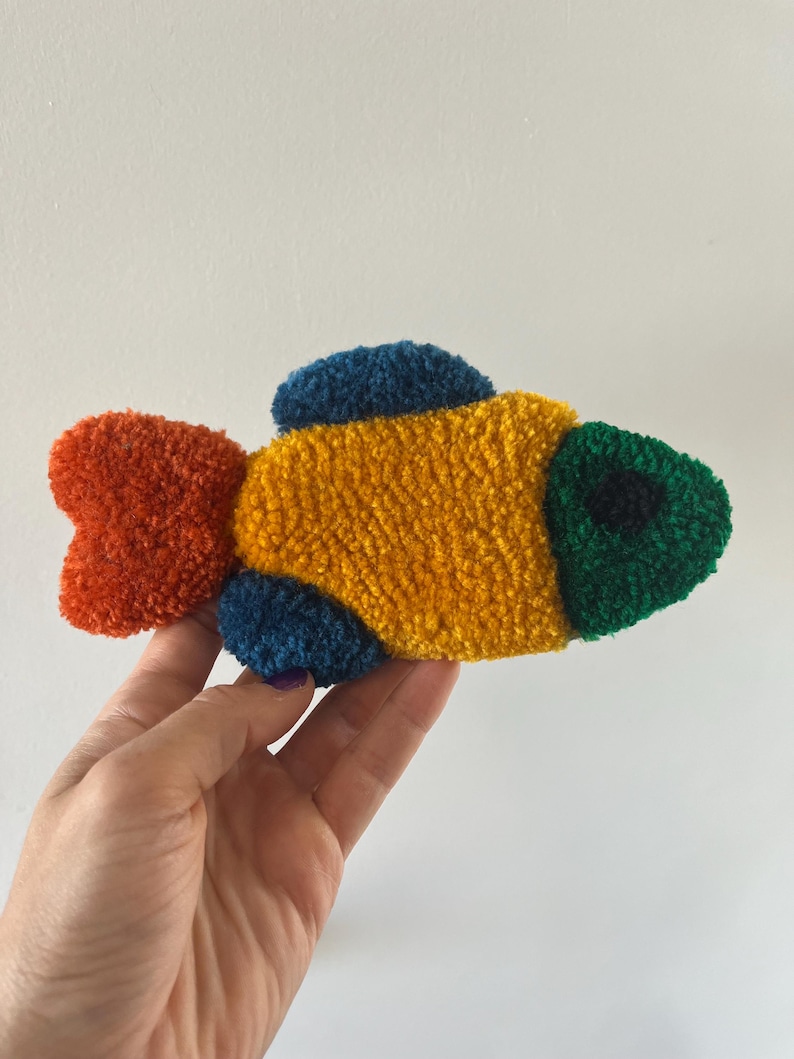 K&ouml;nnte beinhalten: Ein farbenfrohes, strukturiertes Fischornament. Der Fisch hat einen gelben K&ouml;rper, einen roten Schwanz, einen gr&uuml;nen Kopf und blaue Flossen. Das Objekt ist wahrscheinlich handgefertigt und hat ein weiches, pl&uuml;schiges Aussehen.