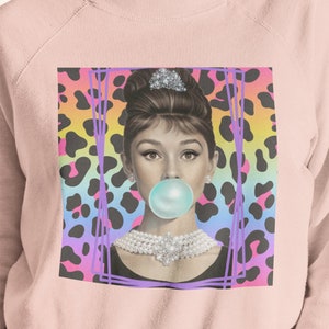 May include: A pink sweatshirt with a graphic print of a woman with a bubblegum in her mouth. The woman is wearing a pearl necklace and a black dress. The background is a rainbow leopard print.