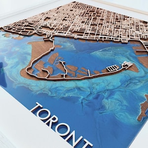 Könnte beinhalten: Eine gerahmte, dreidimensionale Karte von Toronto. Die Karte zeigt eine detaillierte Holzauflage der Straßen und Gebäude der Stadt, vor einem blauen Harz, das das Wasser darstellt. Das Wort "TORONTO" ist in weißen Buchstaben ausgeschnitten.