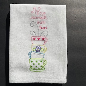 May include: White kitchen towel with embroidered stacked teacups in pink, yellow, blue, and green. The text "My cup runneth over Ps 23:5" is embroidered in pink and gray above the teacups. The towel is made of a textured fabric.