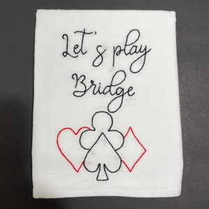 May include: White rectangular kitchen towel with the embroidered text "Let's play Bridge" in black script. Below the text are red and black playing card suit symbols: heart, club, and diamond.