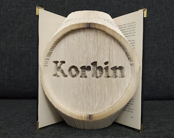 Puede incluir: Un libro doblado en forma de círculo con el nombre "Korbin" recortado en el centro. El libro está abierto y de pie sobre una superficie oscura.