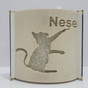 May include: A book with a cat silhouette cut out of the pages. The book is folded into a 3D shape and the name "Nese" is cut out of the top of the book.