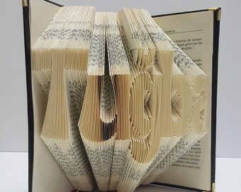 Puede incluir: Una escultura de arte de libro de la palabra "Tiger" creada doblando las páginas de un libro. El libro está abierto y las páginas están dobladas para crear las letras de la palabra. El libro está encuadernado en negro y tiene un acento dorado en el lomo.