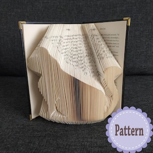 May include: A book folded into the shape of a bird. The book is open and the pages are folded to create the shape of the bird. The book is sitting on a dark grey surface. The text on the book is in German. The image also includes a white circle with the word "Pattern" in black text.
