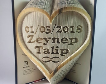 Puede incluir: Un libro con forma de corazón con la fecha 01/03/2015 y los nombres "Zeynep" y "Talip" en su interior. El corazón está hecho de páginas dobladas y tiene un símbolo de infinito en la parte inferior.