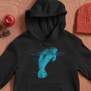 May include: Black hoodie with a blue manatee graphic. The manatee is swimming in water with ripples. The hoodie has a drawstring hood and a kangaroo pocket.