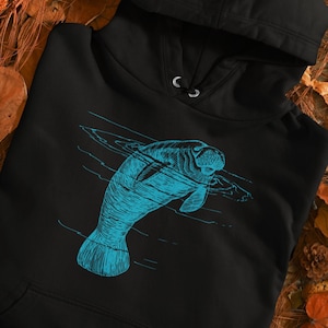 Könnte beinhalten: Schwarzer Hoodie mit einer blauen Umrandung eines Seekuhs, der im Wasser schwimmt. Die Seekuh blickt nach rechts.
