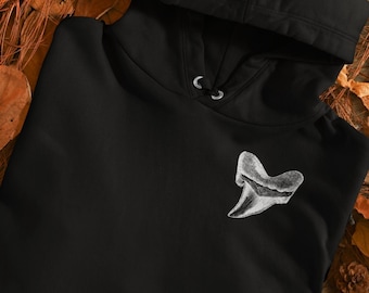 Puede incluir: Sudadera con capucha negra con un diseño de diente de tiburón bordado en blanco. La sudadera está colocada sobre un lecho de hojas marrones.