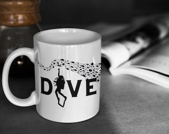 Puede incluir: Taza de cerámica blanca con una silueta negra de un buceador y la palabra "DIVE" impresa en ella. El buceador está rodeado por un rastro de pequeños peces negros.