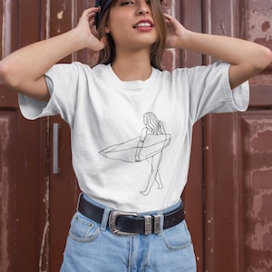 May include: A white t-shirt with a line drawing of a person surfing. The person is wearing a swimsuit and holding a surfboard. The t-shirt is being worn by a person with brown hair and a black cap. The person is wearing blue denim shorts and a black belt.