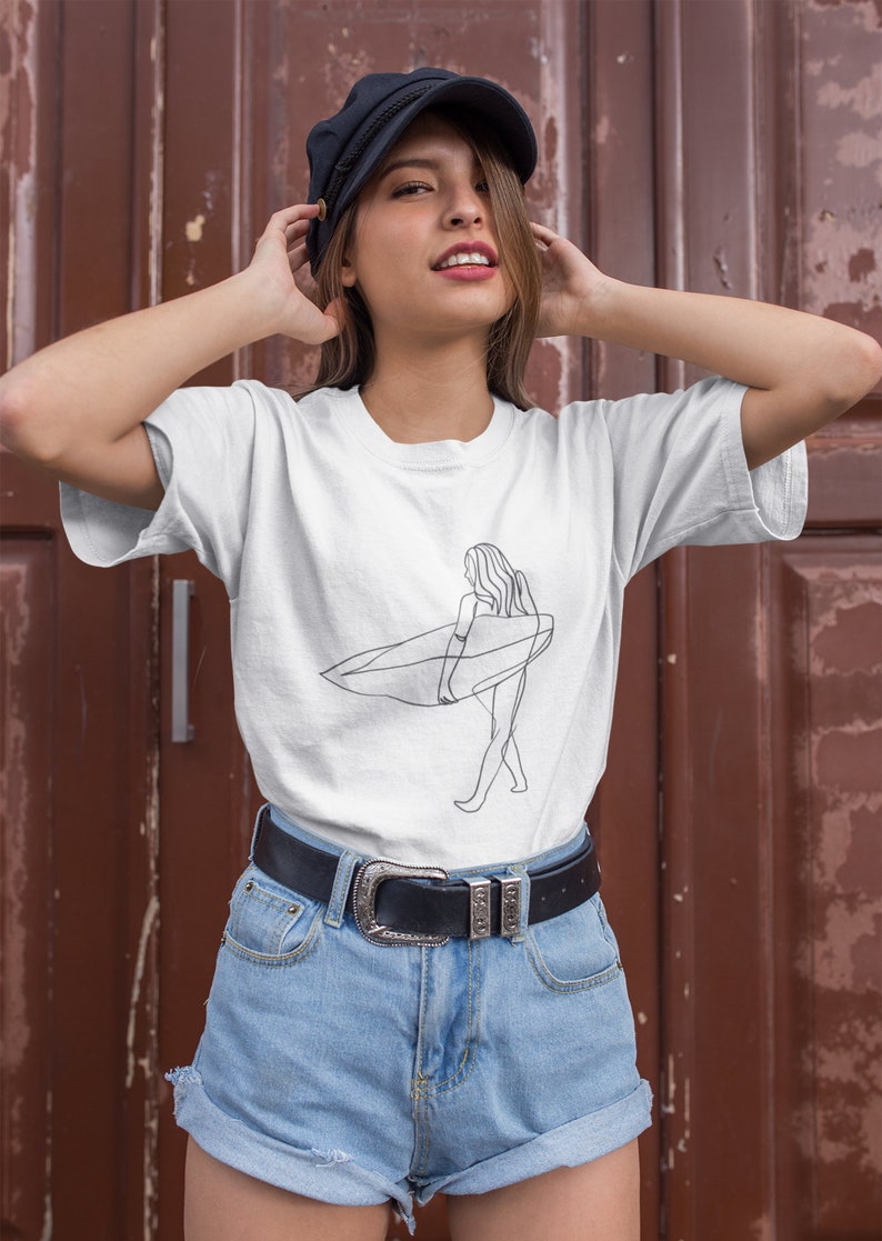 May include: A white t-shirt with a line drawing of a person surfing. The person is wearing a swimsuit and holding a surfboard. The t-shirt is being worn by a person with brown hair and a black cap. The person is wearing blue denim shorts and a black belt.