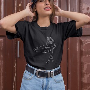 May include: A black t-shirt with a white line drawing of a person surfing. The person is holding a surfboard and walking on the beach. The t-shirt is being worn by a person with brown hair and a black cap.